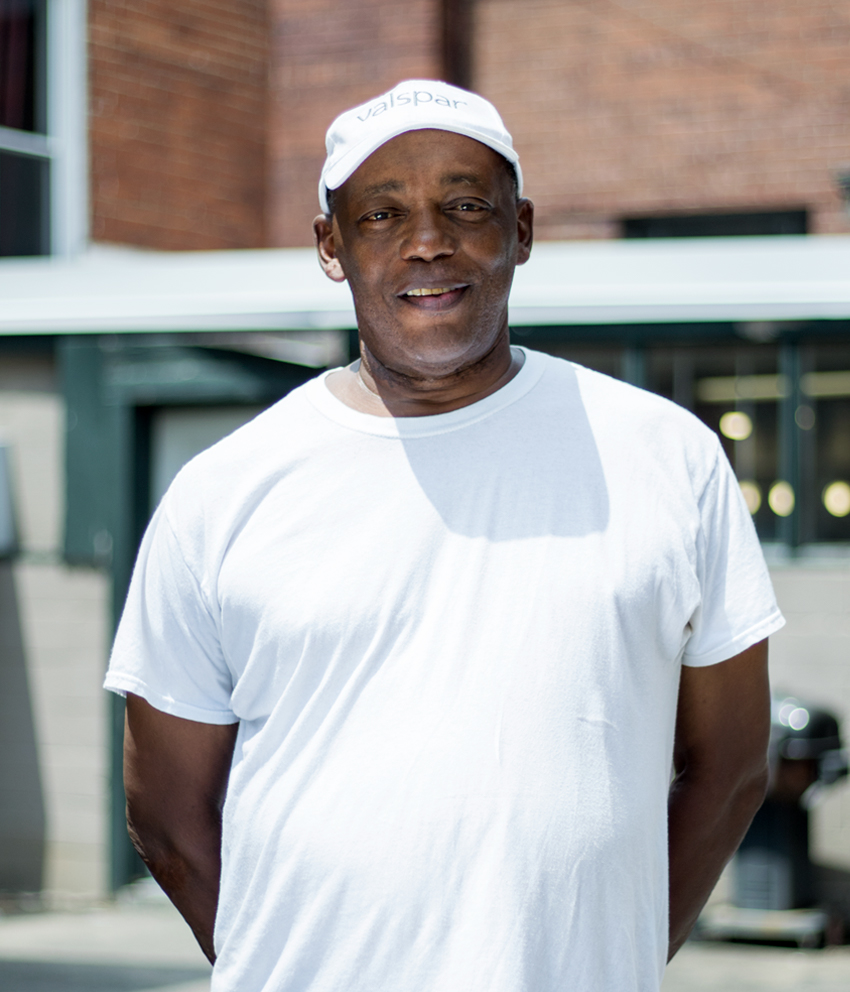 A man smiling, wearing a Valspar branded cap and white t-shirt, with his hands behind his back