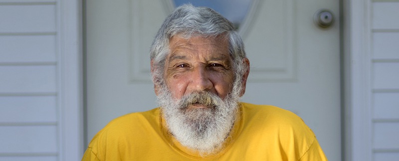 Portrait of an elderly man with wrinkles on his forehead and facial hair including a mustache, beard, and hair, wearing a yellow t-shirt, sitting in front of a wooden door.