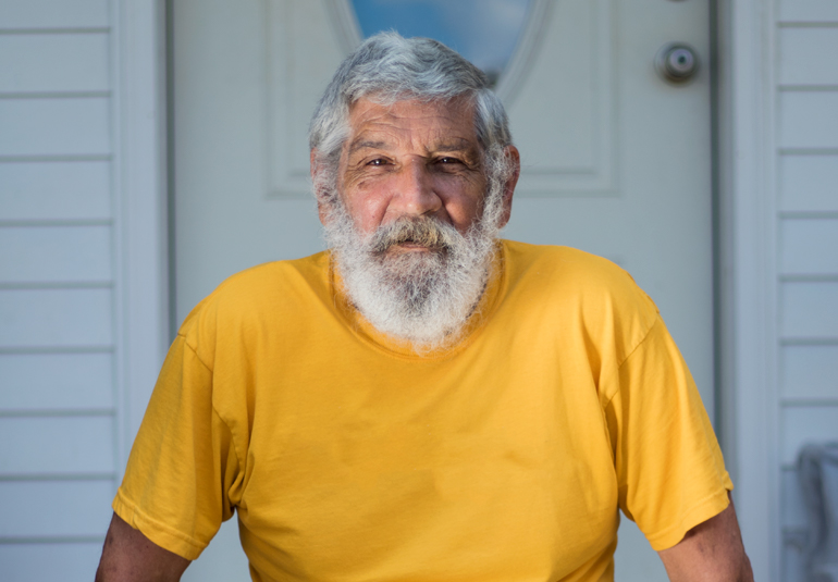 Close up portrait of an elderly man with wrinkles on his forehead and facial hair including a mustache, beard, and hair, wearing a yellow t-shirt, sitting in front of a wooden door.