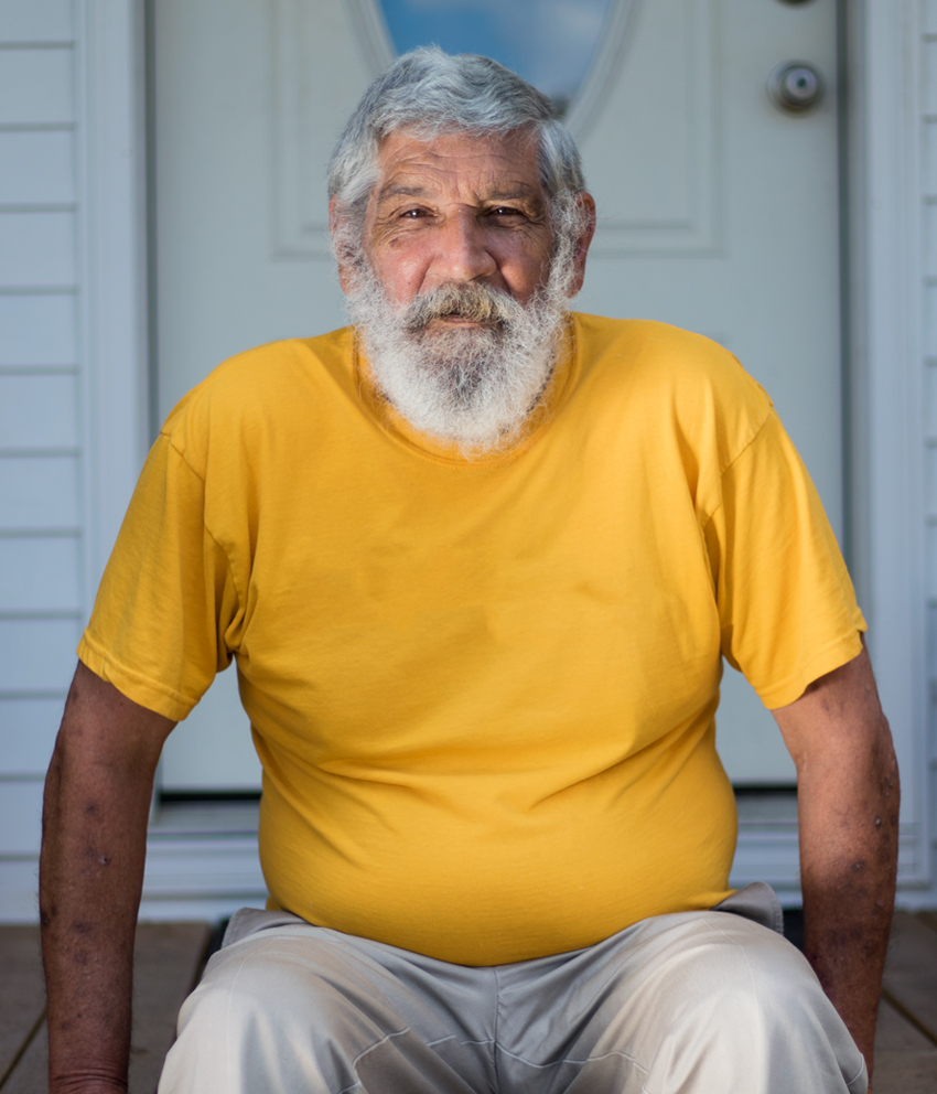 Portrait of an elderly man with wrinkles on his forehead and facial hair including a mustache, beard, and hair, wearing a yellow t-shirt, sitting in front of a wooden door.